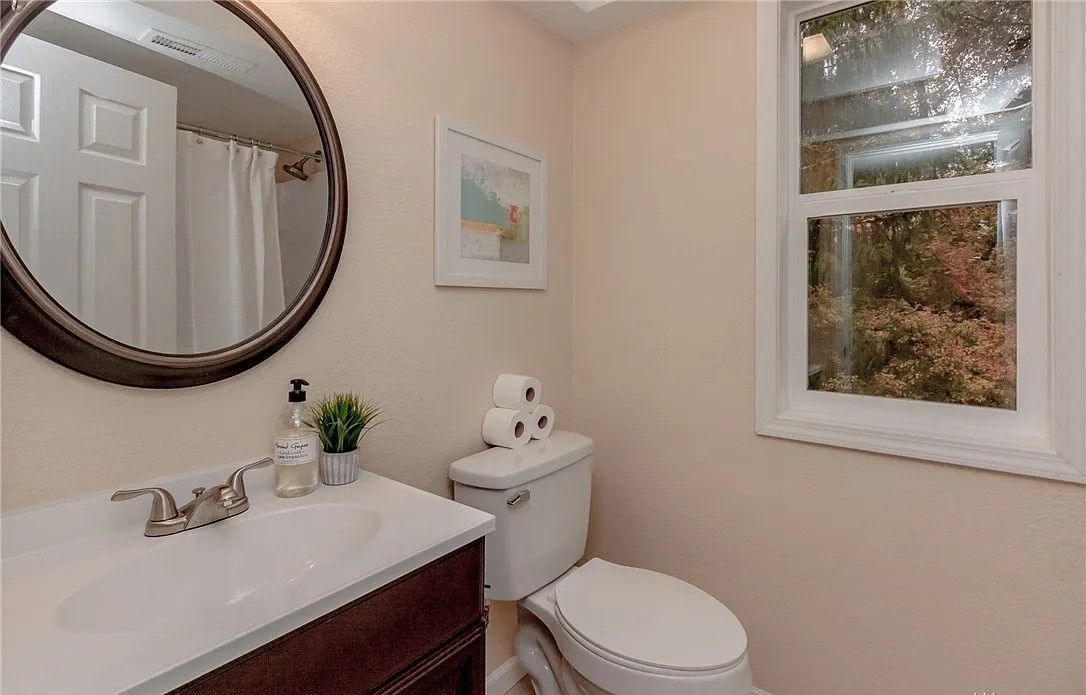 Bathroom with a dark vanity, round mirror, white toilet, and window with outdoor view.