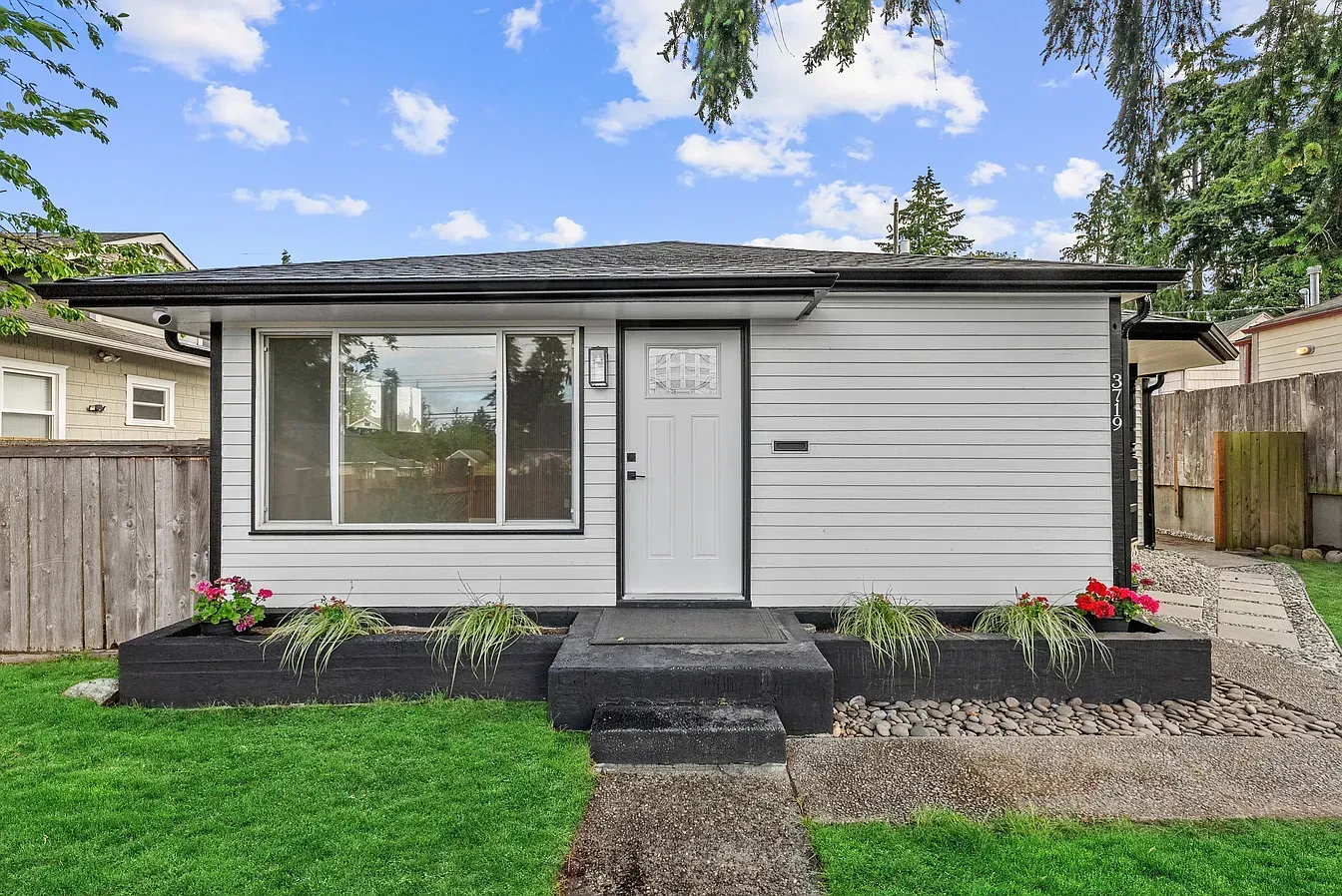 Small gray house with white door, front window, black steps, and flower beds in a fenced yard.