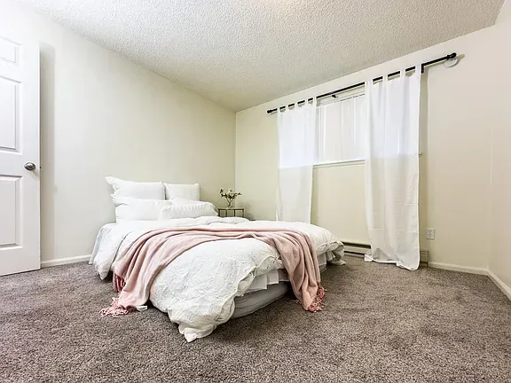 Bedroom with white walls, a bed with white bedding and a pink throw, and white curtains.