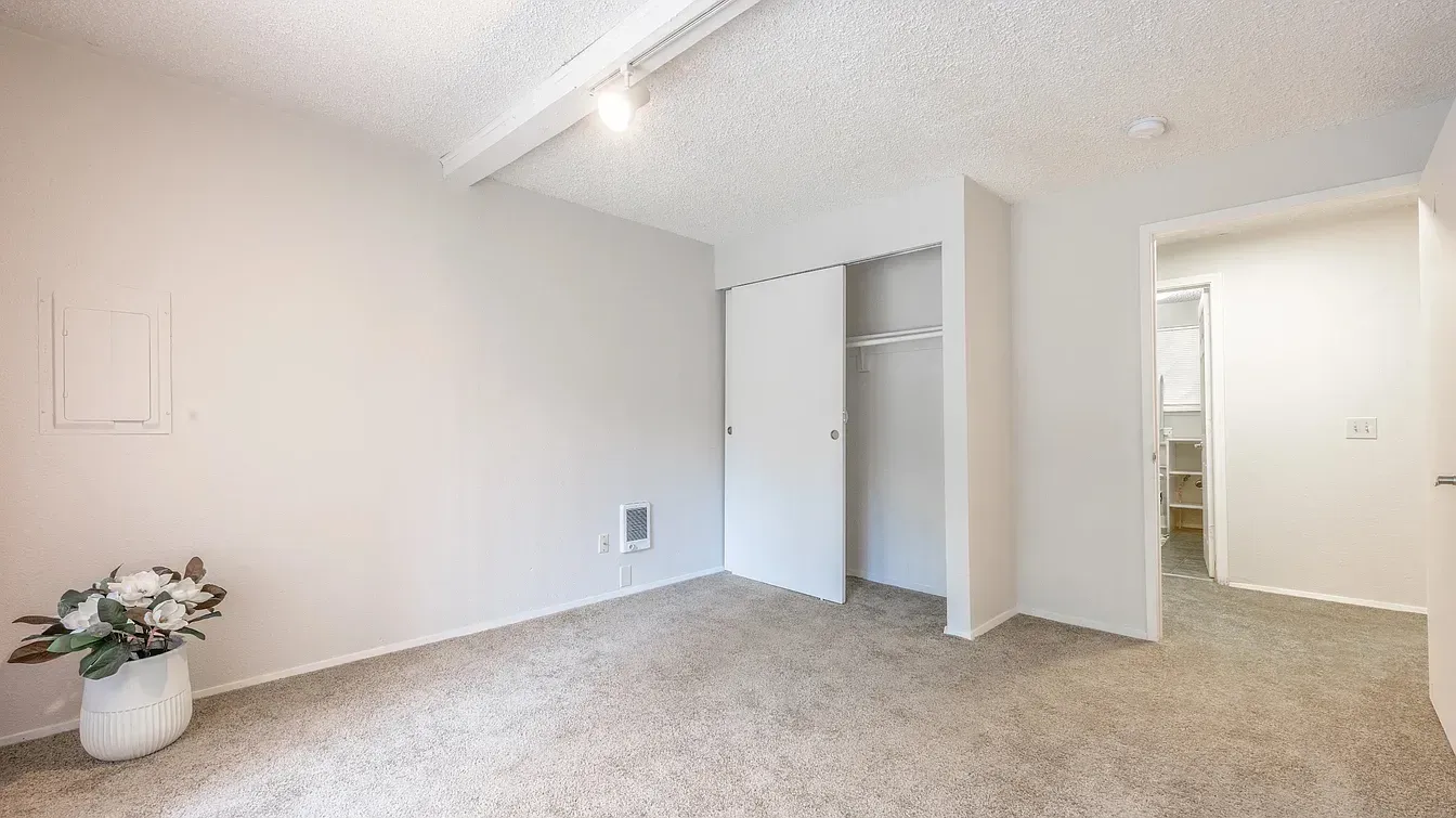 Empty bedroom with a closet and a doorway to a bathroom. Light tan carpet and white walls.