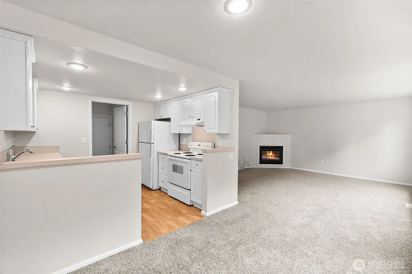 Bright open living room with carpet, white kitchen, and a small fireplace on the far wall