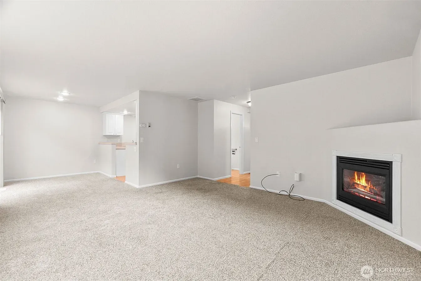 Empty carpeted living room with white walls, gray fireplace, and a small kitchen area in the back
