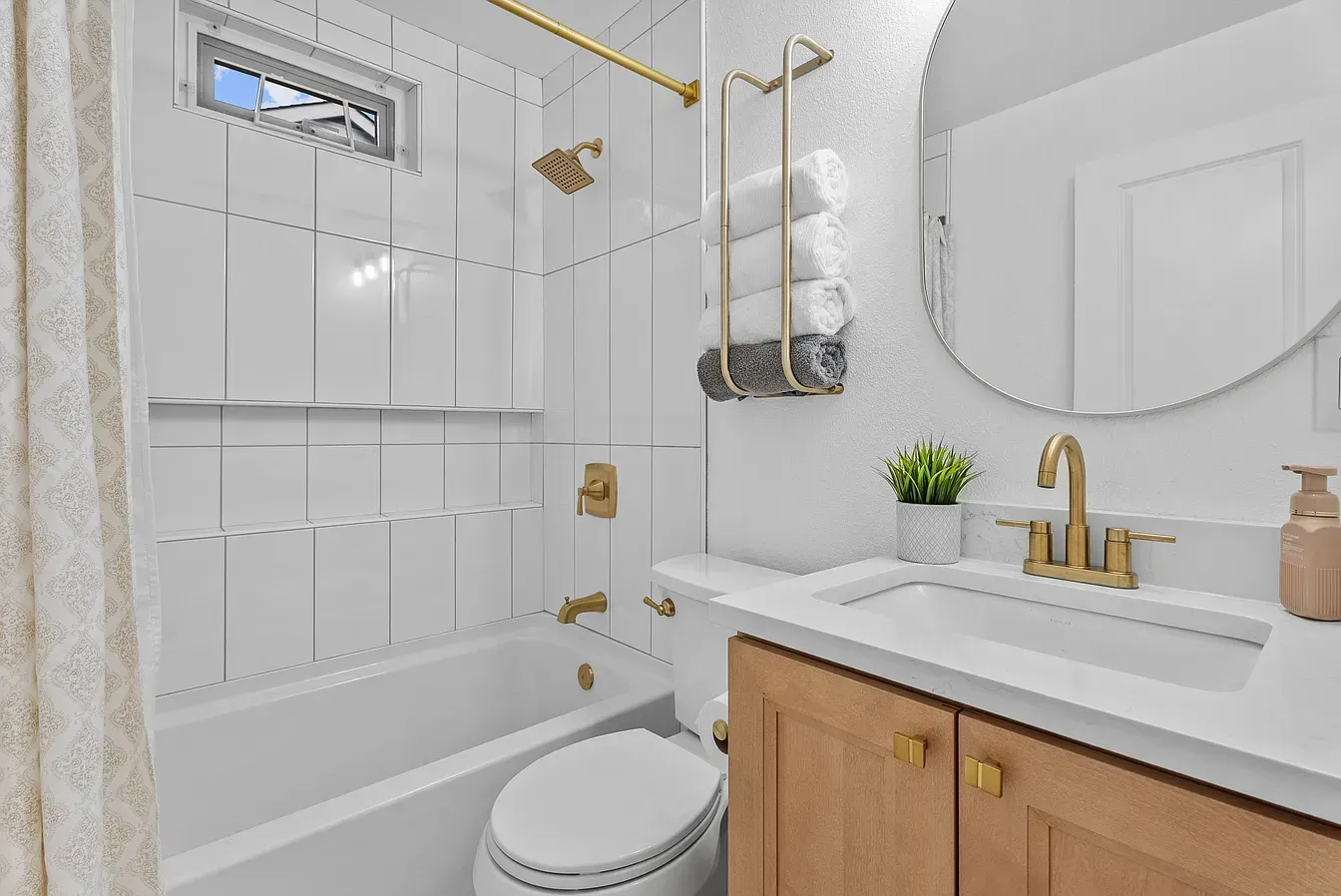 Bright white bathroom with tub-shower, gold fixtures, vanity, round mirror, and wood cabinets