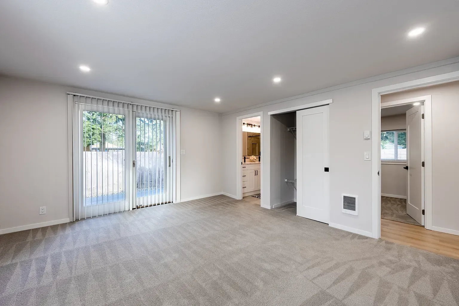 Empty carpeted living room with sliding glass doors, recessed lights, and open doorways to adjacent rooms