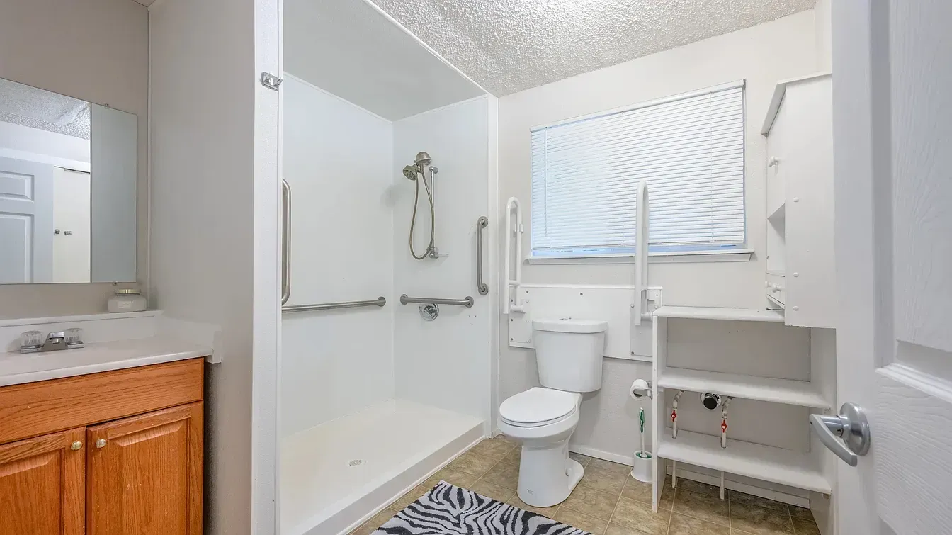 Accessible bathroom with walk-in shower, grab bars, open shelving, and toilet near a window with blinds.