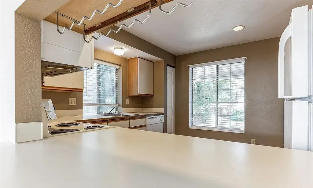Kitchen with a white countertop, beige walls, and light wooden cabinets. A window and refrigerator are also visible.