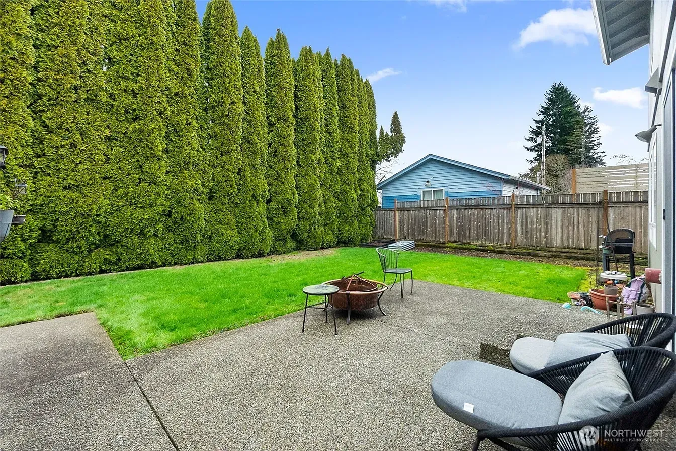 Backyard patio with gravel, lawn, tall hedge, fire pit, and outdoor seating beneath a blue sky