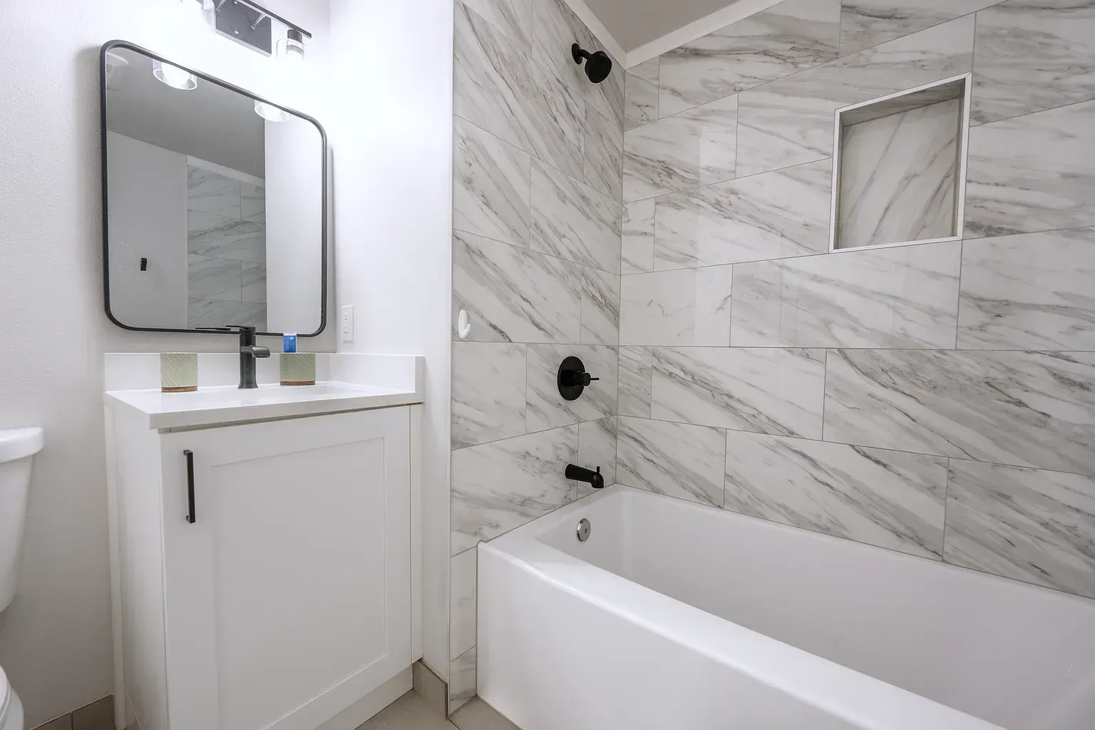 Bright white bathroom with vanity, mirror, and marble-tiled tub surround