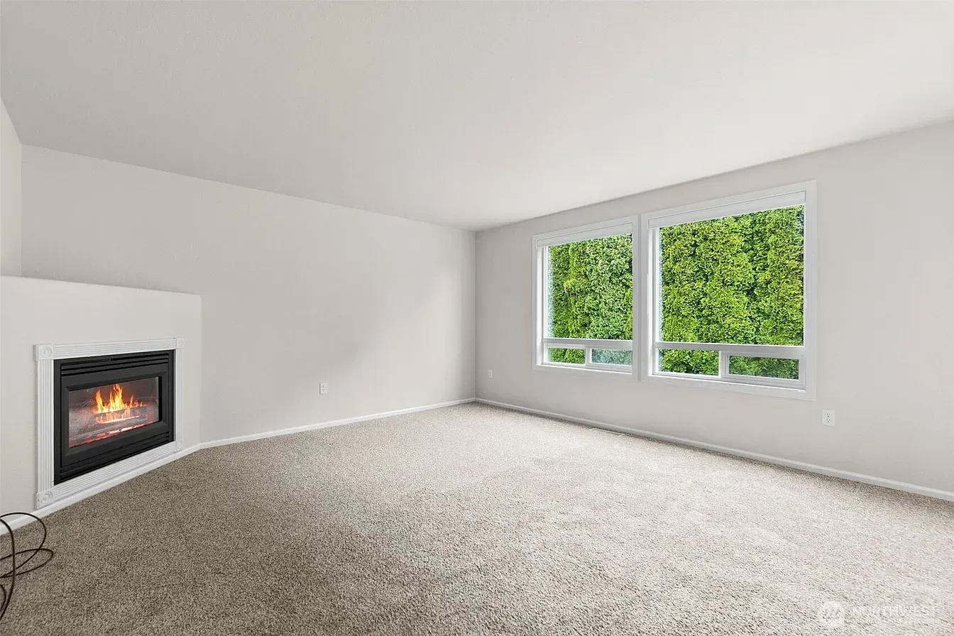 Empty white living room with a modern fireplace, carpeted floor, and large windows overlooking green trees