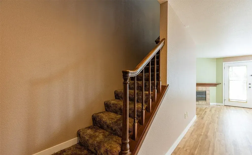 Staircase with brown carpet and wooden railing. Interior shot, adjacent to a room with fireplace and door.