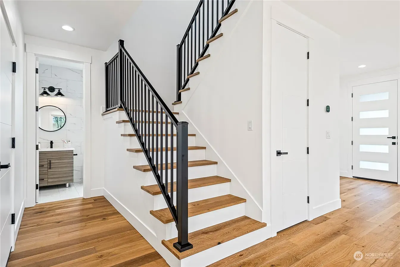 Wooden stairs with black railing in a bright white hallway with a bathroom and front door.