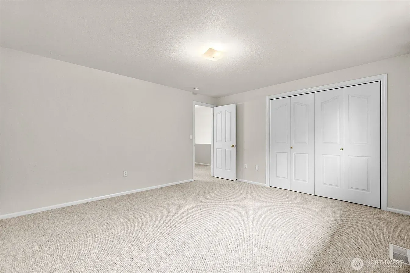 Empty beige bedroom with carpet, white walls, ceiling light, and closet doors open near the entry.