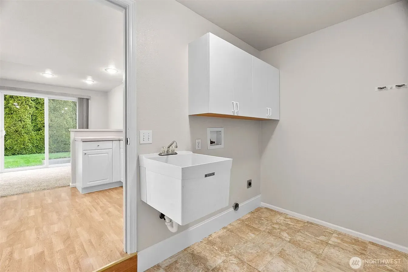 Small laundry room with white cabinets and sink, beige carpet, and wood flooring leading to a bright adjacent room