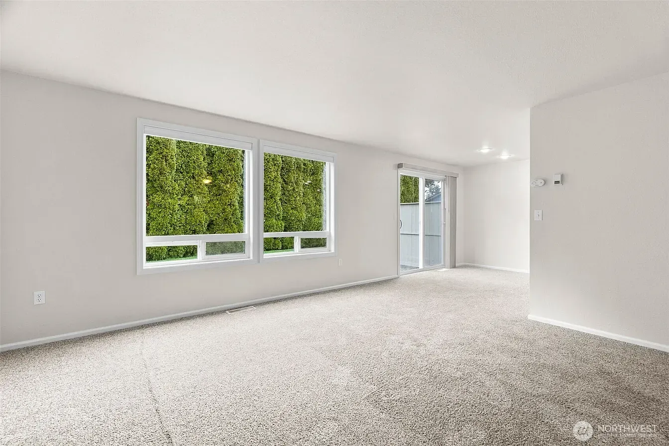 Bright empty room with white walls, carpeted floor, large window, and doorway to a balcony.