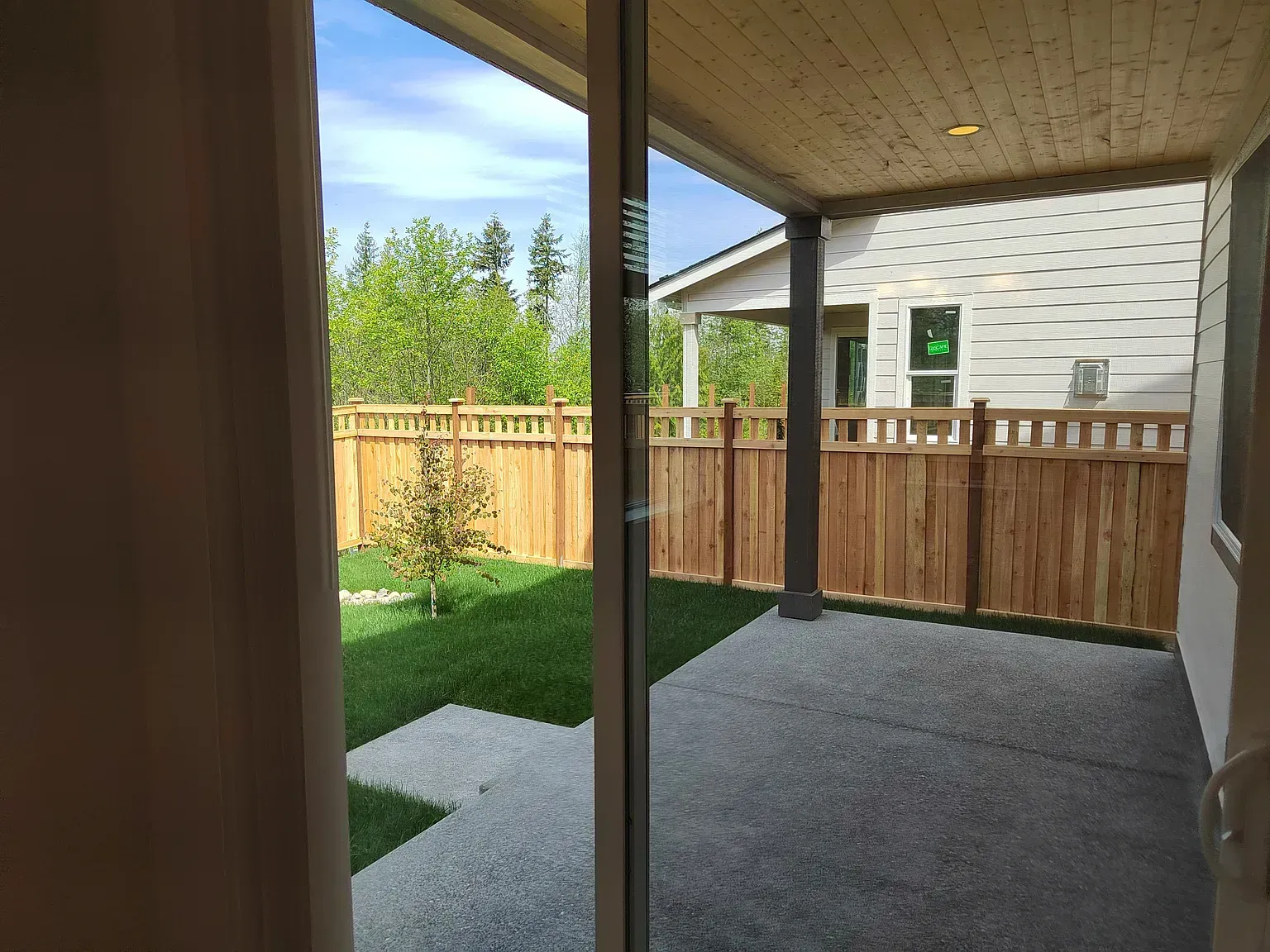View from a doorway: backyard with grass, wooden fence, patio, and a house in the distance.