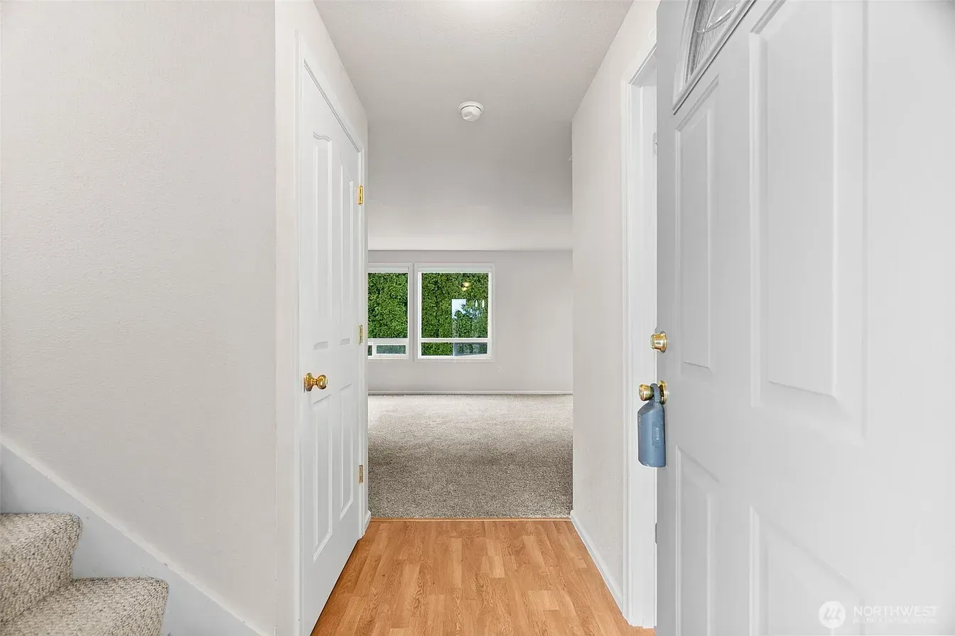 Bright hallway with white doors, wood floor, and a carpeted room with a window at the end.