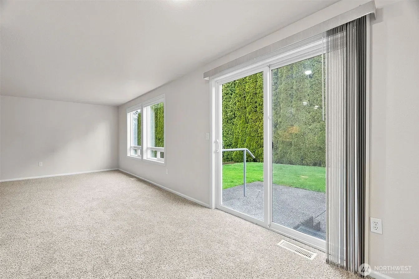 Empty bright living room with white walls, carpeted floor, and sliding glass doors to a green yard