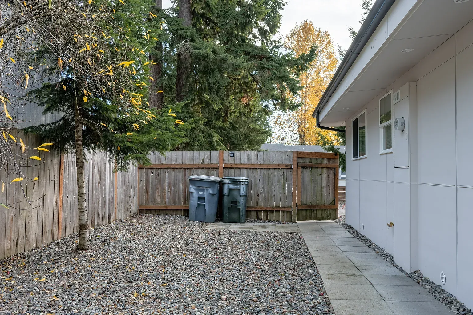 Backyard with gravel, fence, trash bins, and the side of a white house
