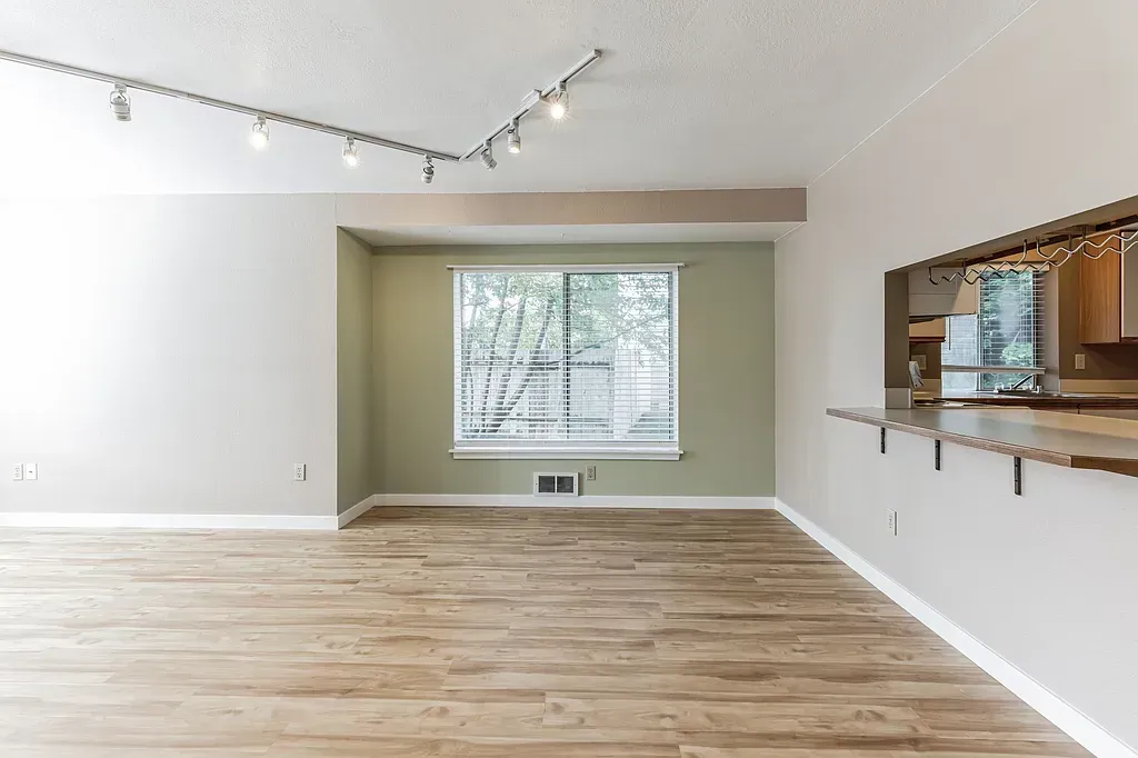 Empty room with light wood floors, a window, and a pass-through to the kitchen.
