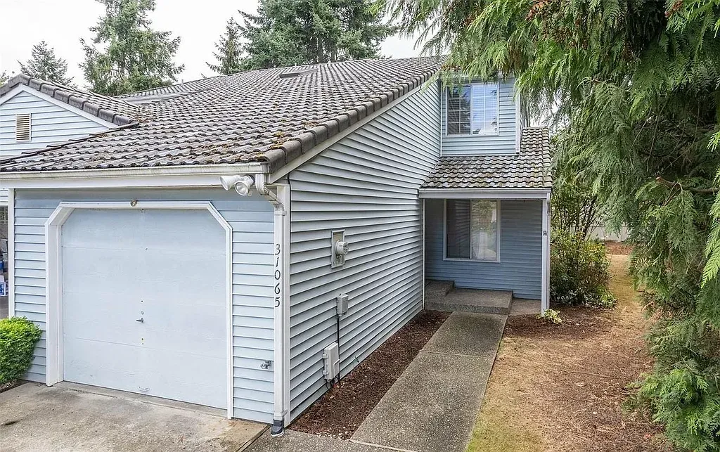 Light blue house with a garage and walkway. Brown roof, surrounded by trees and dry grass.
