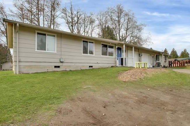 Low-slung, light-colored building with multiple windows, door, and green lawn. Cloudy blue sky and dirt driveway in front.