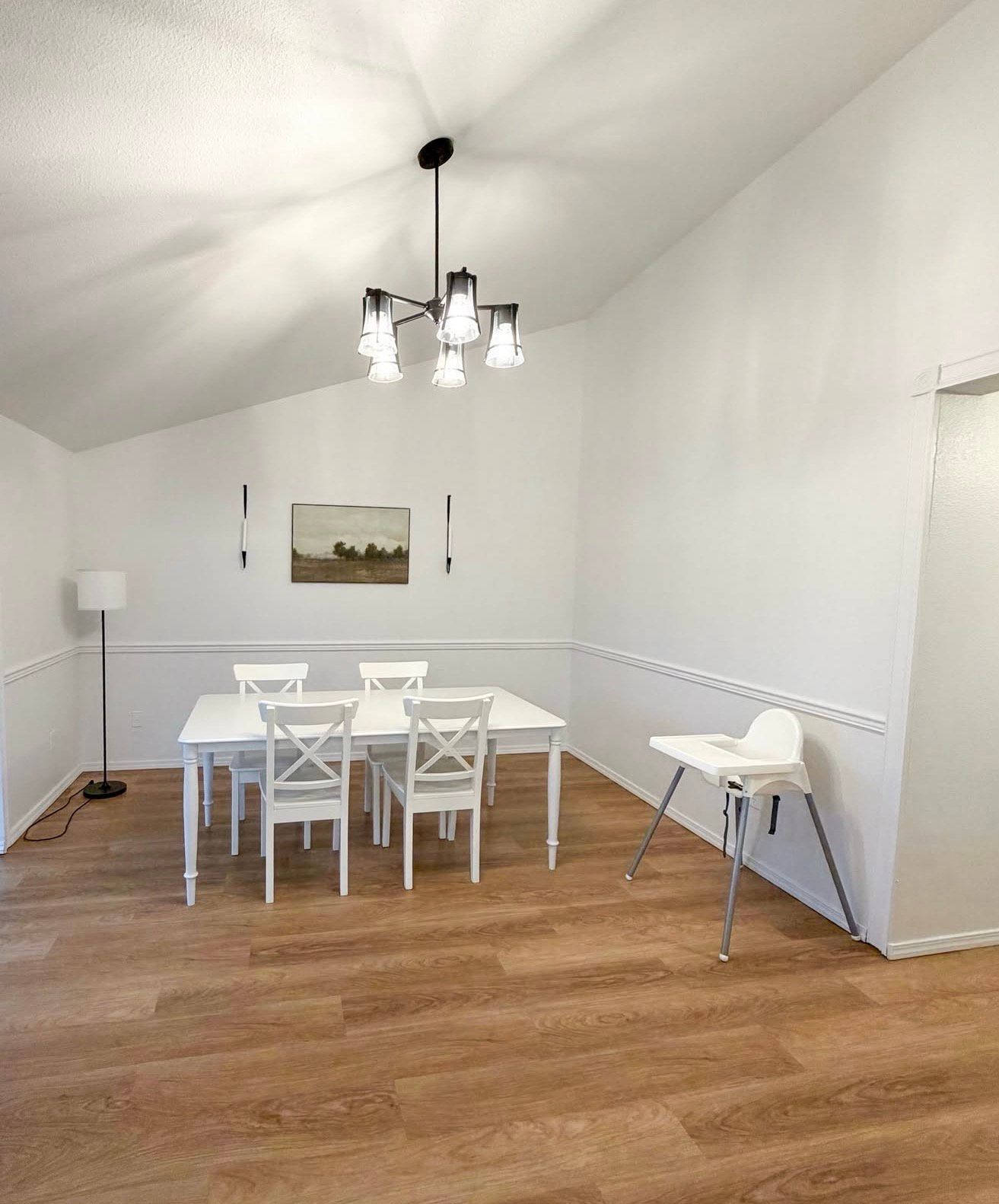 Dining room with white table and chairs, chandelier, high chair, artwork, and wood-look flooring.