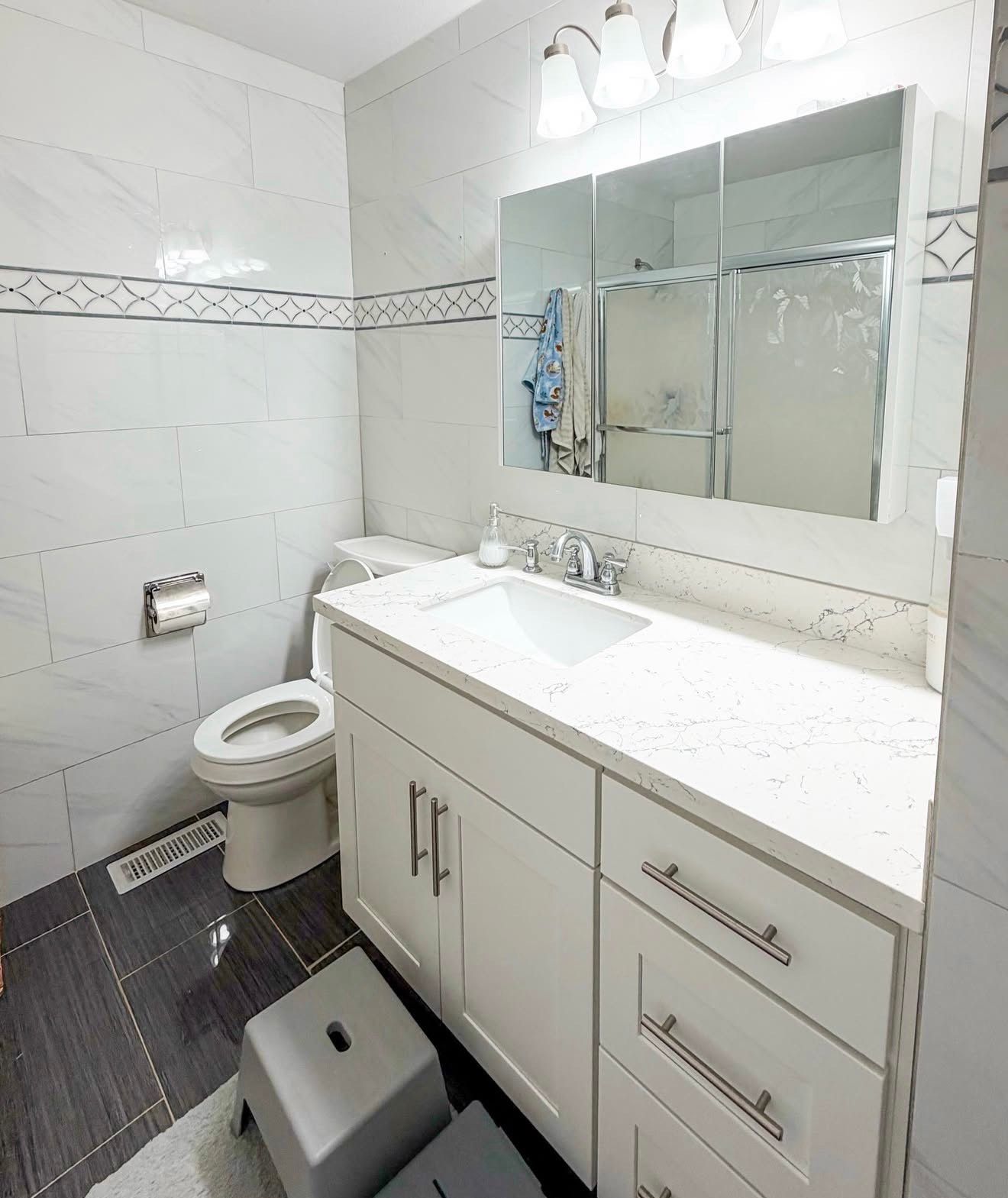 White bathroom with a vanity, toilet, and shower. Gray and white tile, a mirror, and a step stool are visible.