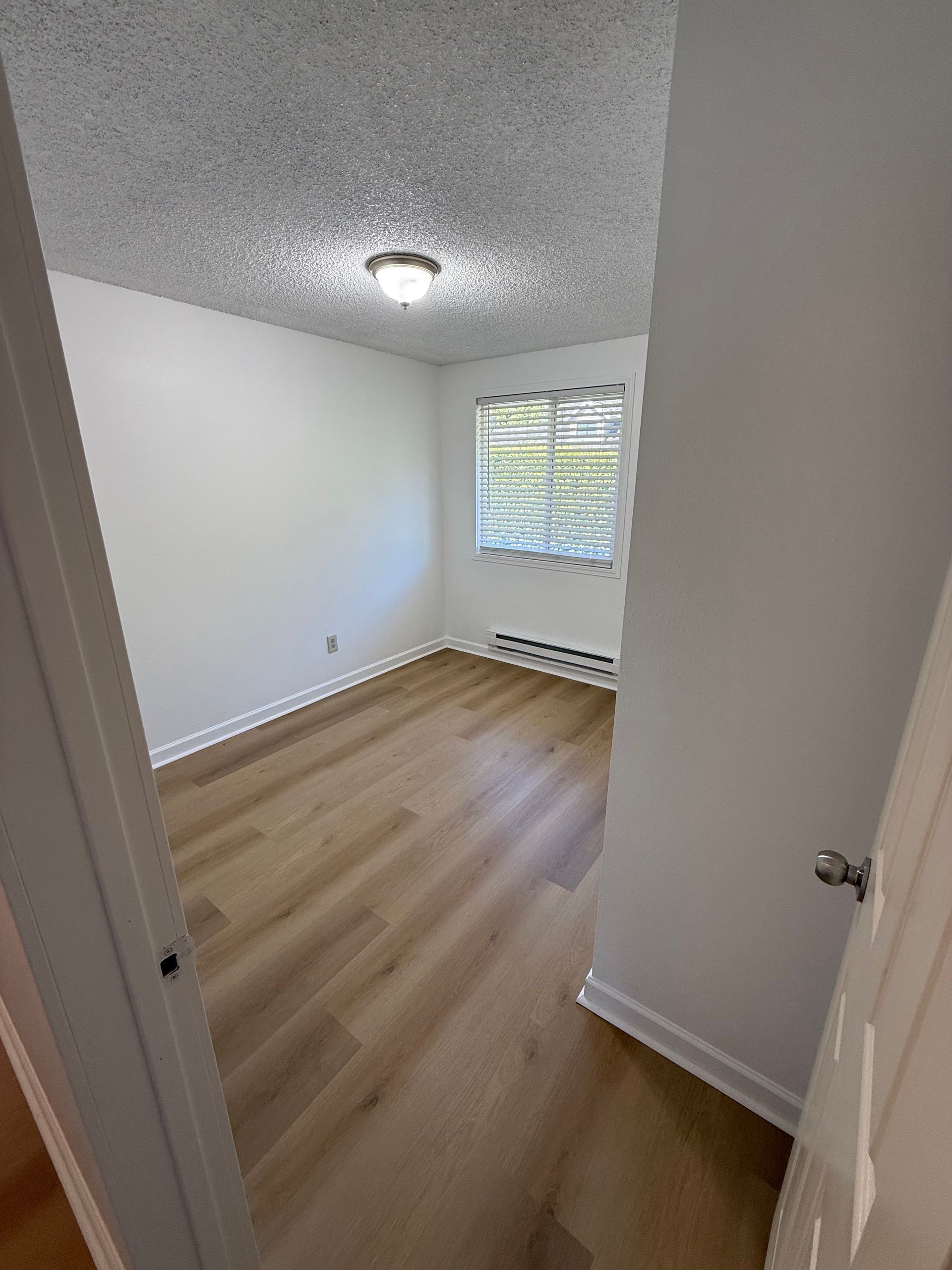 Empty room with light wood flooring, white walls, and a window with a radiator below.