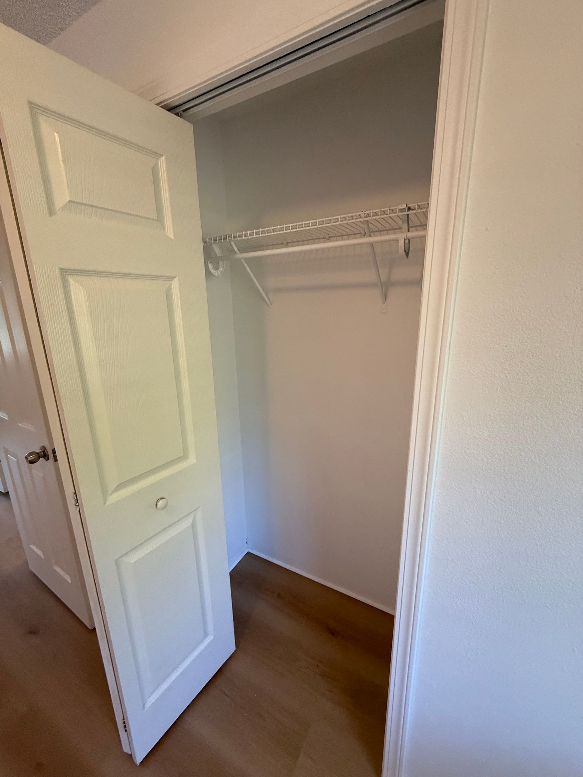 Empty white closet with open door, wire shelf, and wooden floor.