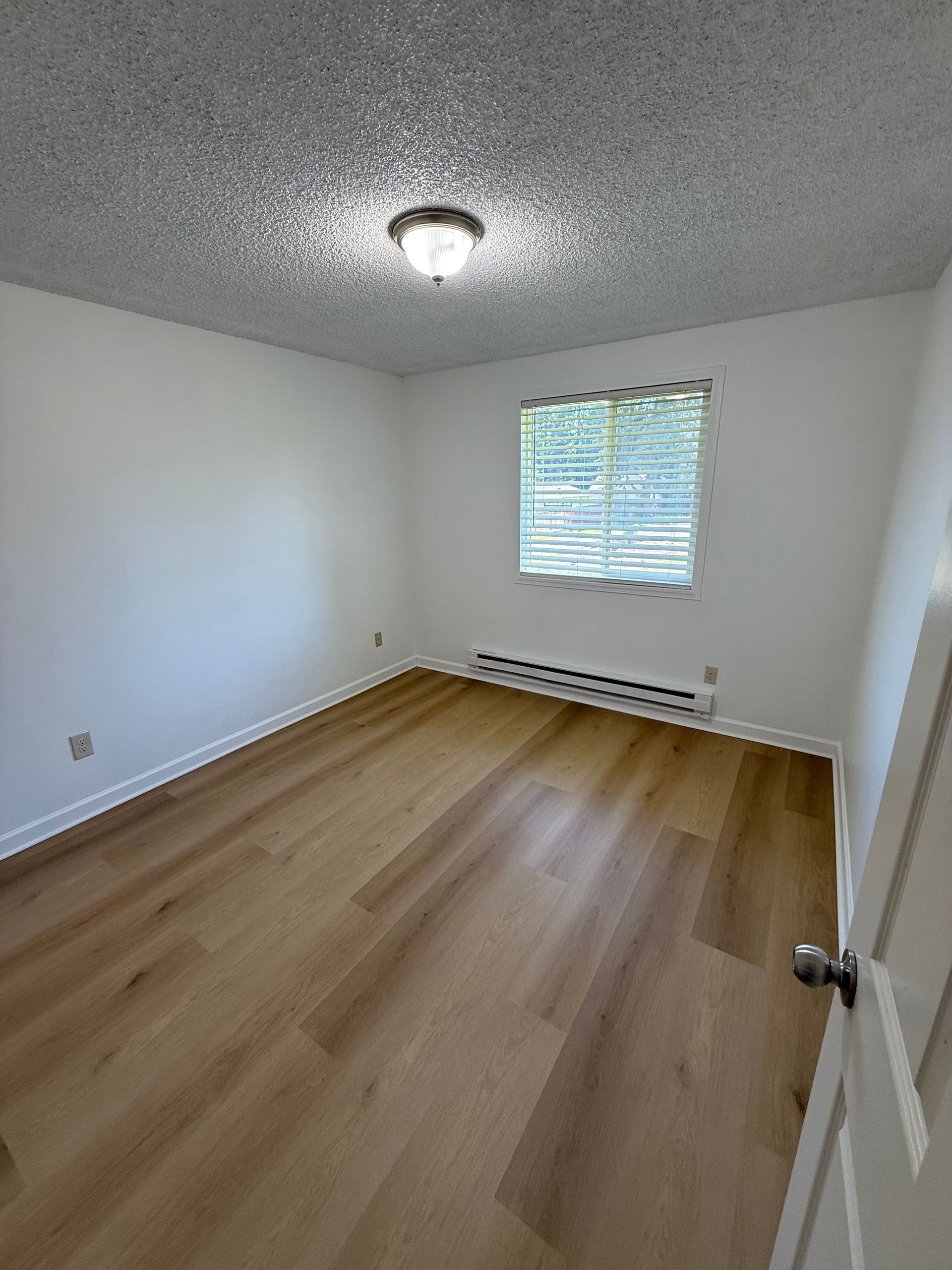 Empty bedroom with light wood flooring, white walls, and a window with blinds.