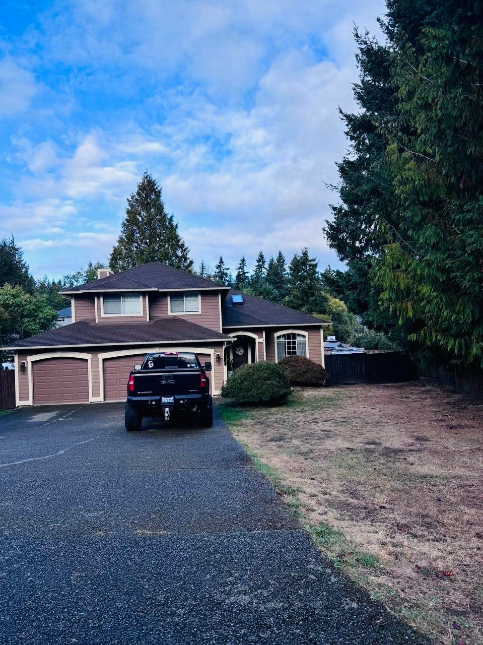 Two-story brown house with a dark pickup truck parked in the driveway; blue sky and trees in the background.