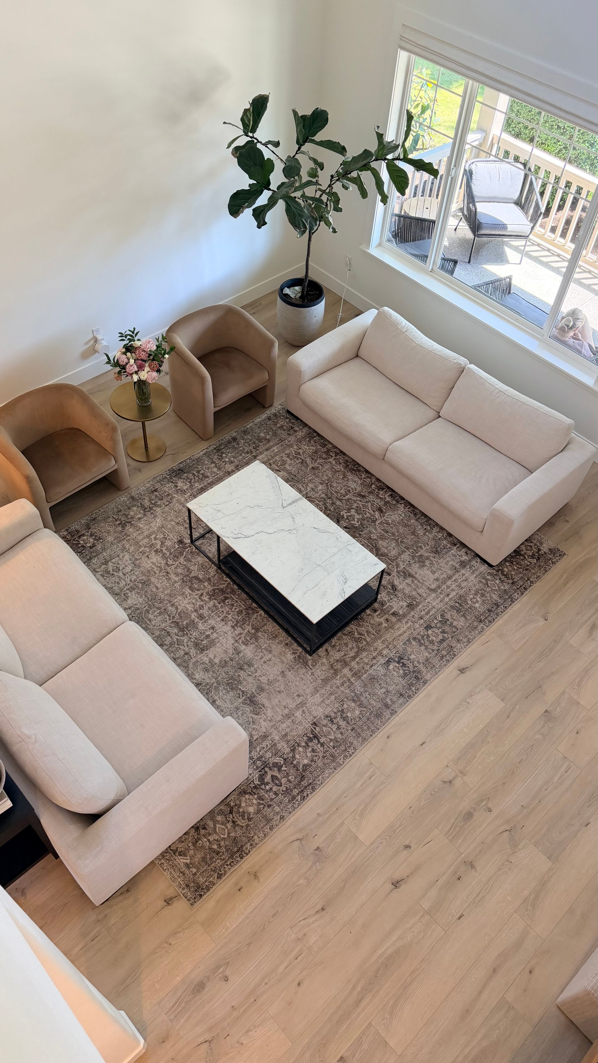 Top-down view of a bright living room with beige sofas, a coffee table, rug, and large window.