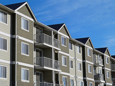 Multi-story apartment building with balconies, olive green and beige siding, blue sky.