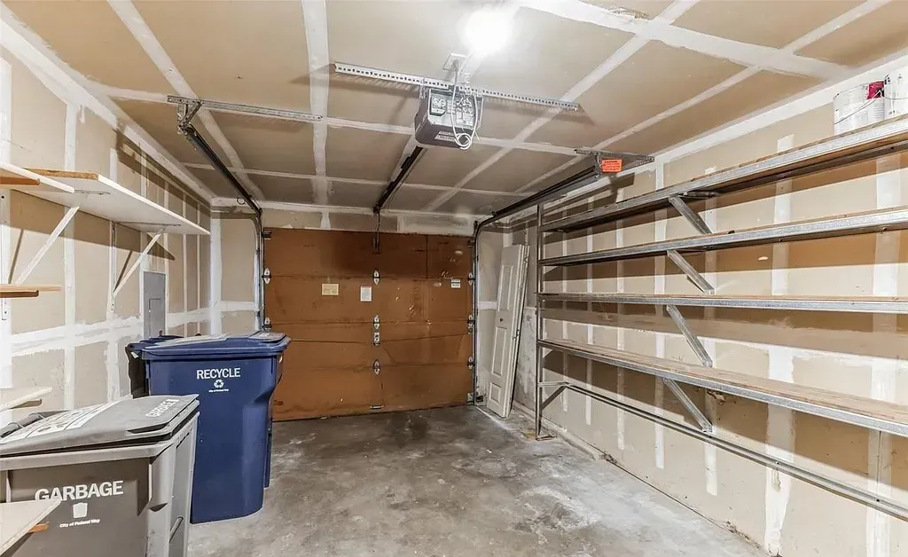 Interior view of a garage with a closed door, shelves, and trash cans. The walls are unfinished.
