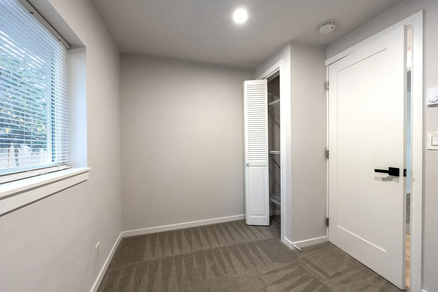 Empty carpeted room with white walls, window blinds, recessed light, and an open closet beside a closed door