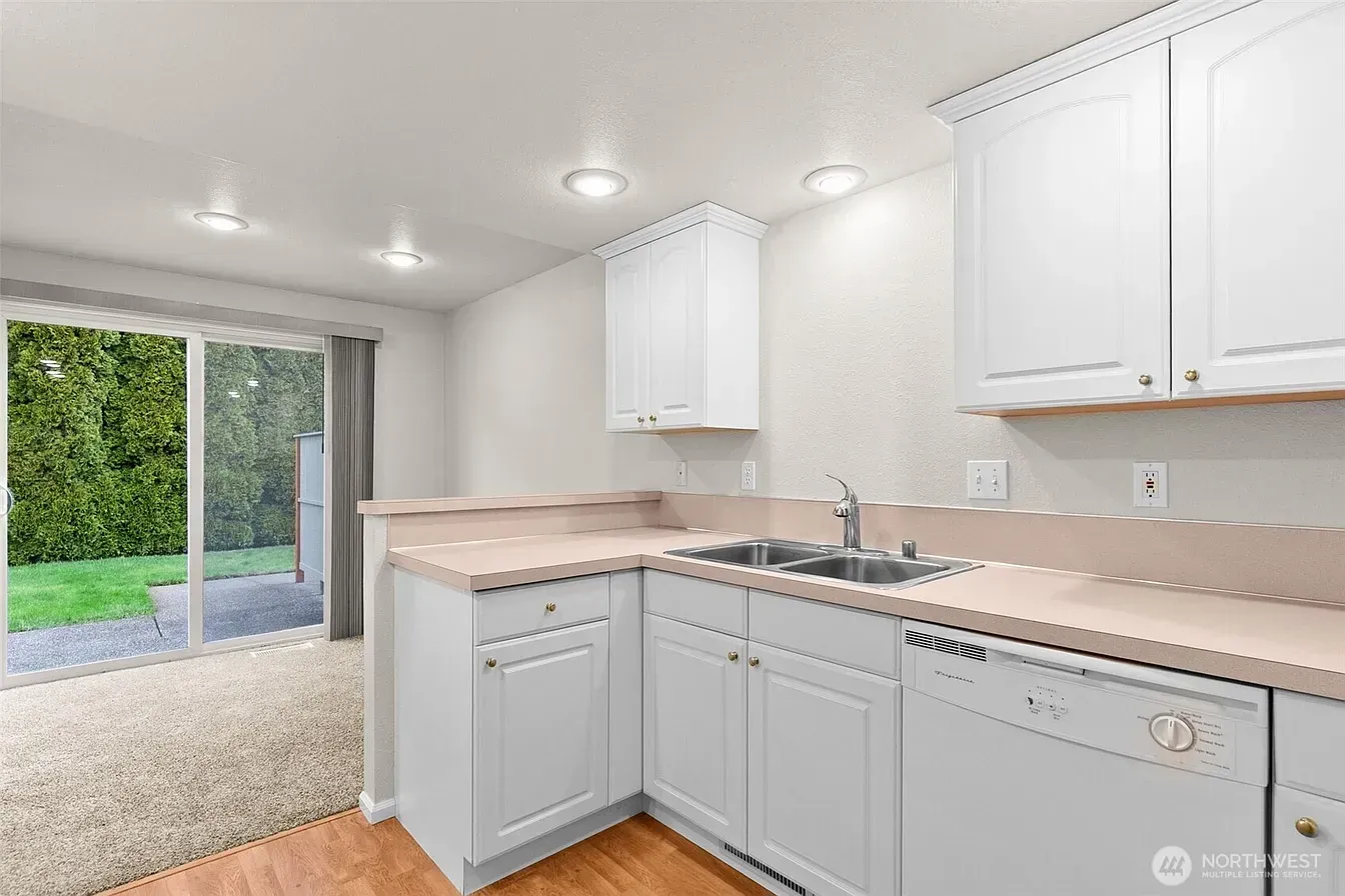 Bright white kitchen with tan countertops, dishwasher, and sliding glass doors to a green yard