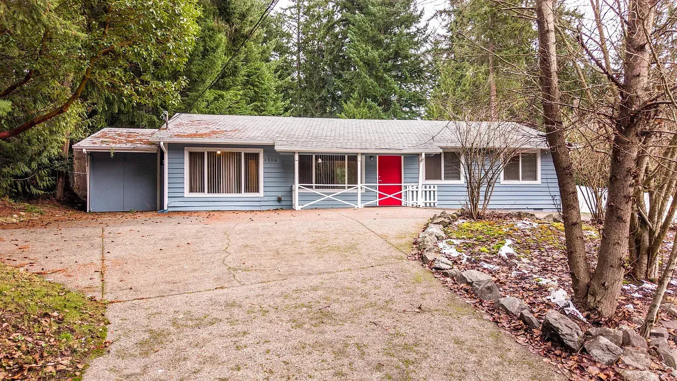 Light blue ranch-style house with red front door, surrounded by trees; driveway in front.