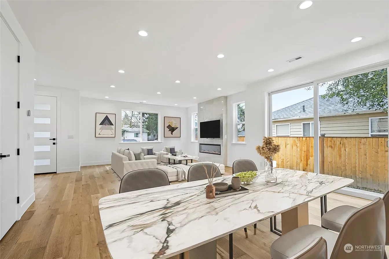 Open-concept living space with dining table, fireplace, and large windows. Bright, modern interior with hardwood floors.
