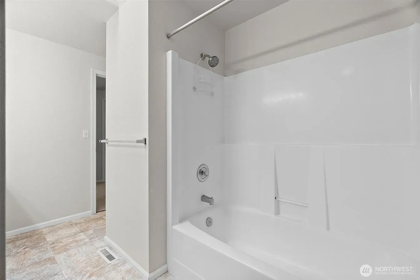 Bright white bathroom with tub, shower fixtures, glass door, and tiled floor leading to a doorway.