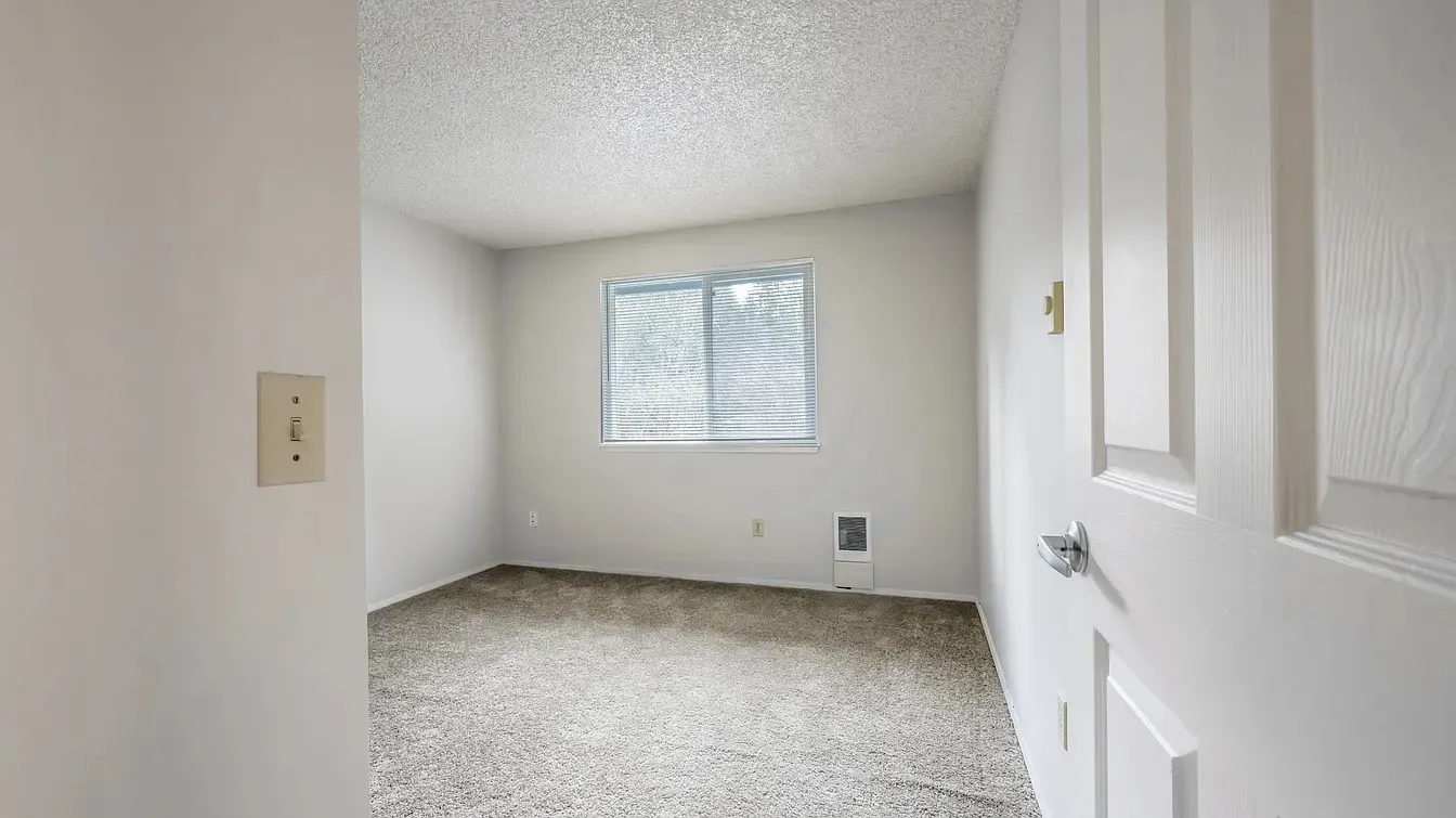 Empty, white-walled room with a window covered by blinds, carpeted floor, and a closed door.