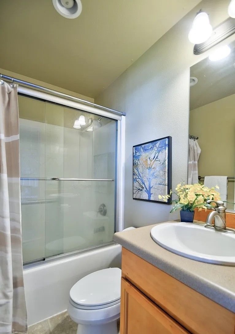 Bathroom with a glass shower, toilet, sink, and artwork. Beige walls and light wood cabinets.