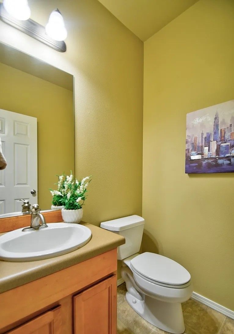 A small bathroom with a toilet, sink, mirror, and a cityscape painting on a yellow wall.