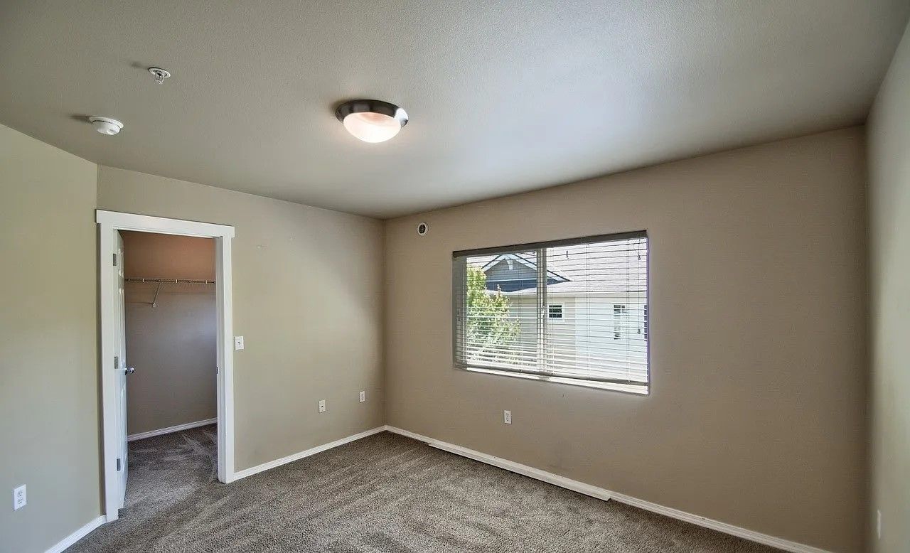 Empty bedroom with beige walls, carpet, window, and a door to a walk-in closet.