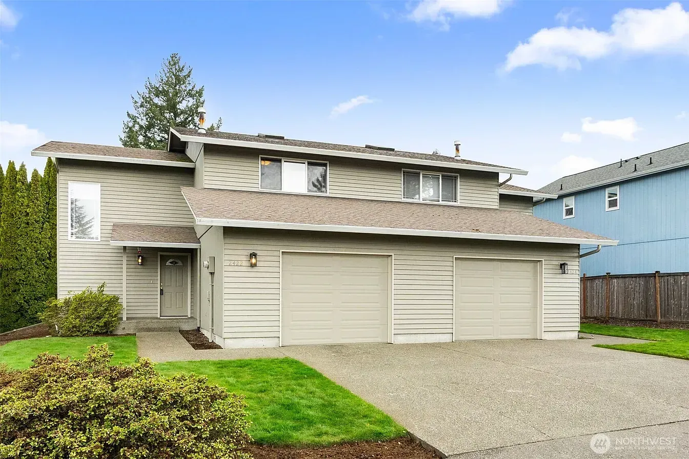 Two-story suburban house with light siding, double garage, and gravel driveway under a partly cloudy sky