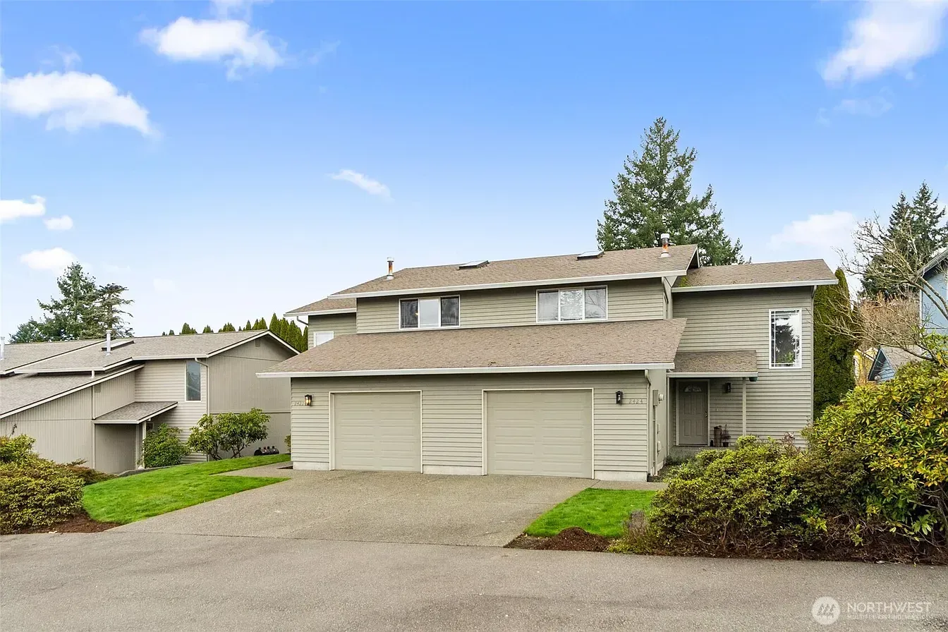 Two-story beige suburban house with a double garage, driveway, and landscaped front yard under a blue sky