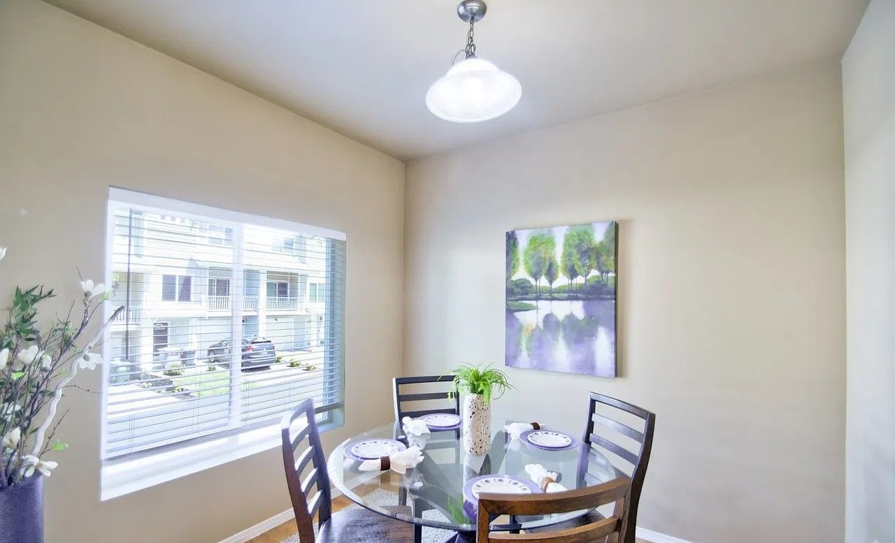 Dining room with a glass-top table, four chairs, a window, and artwork. Neutral walls, light fixture.