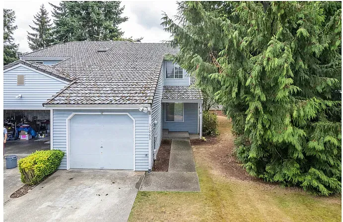 Light blue townhome with a garage, wooden roof, and walkway. Large evergreen tree next to the house.
