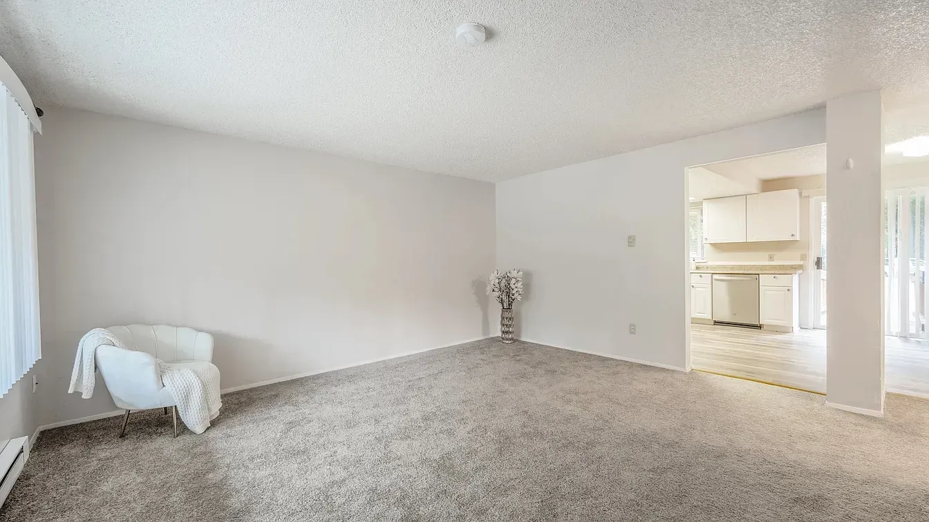 Empty living room with white walls, gray carpet, chair, and view to the kitchen.