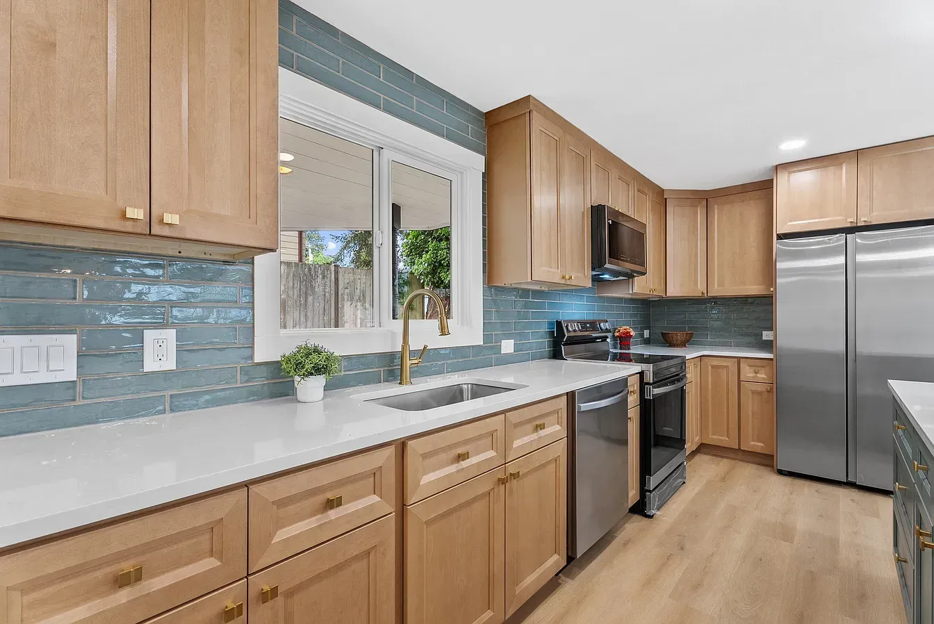 Modern kitchen with light wood cabinets, stainless steel appliances, and a large window over the sink.
