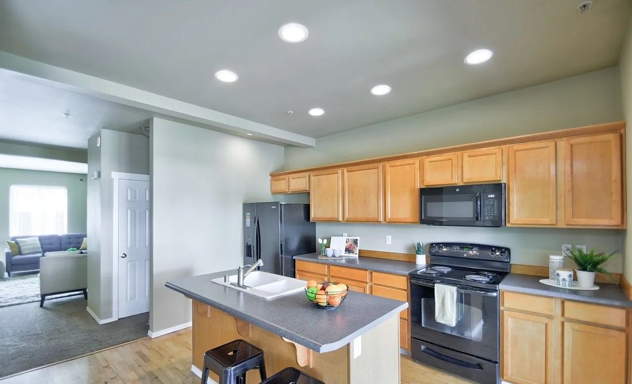 Kitchen with wooden cabinets, black appliances, and island with sink.  Living room visible in the background.